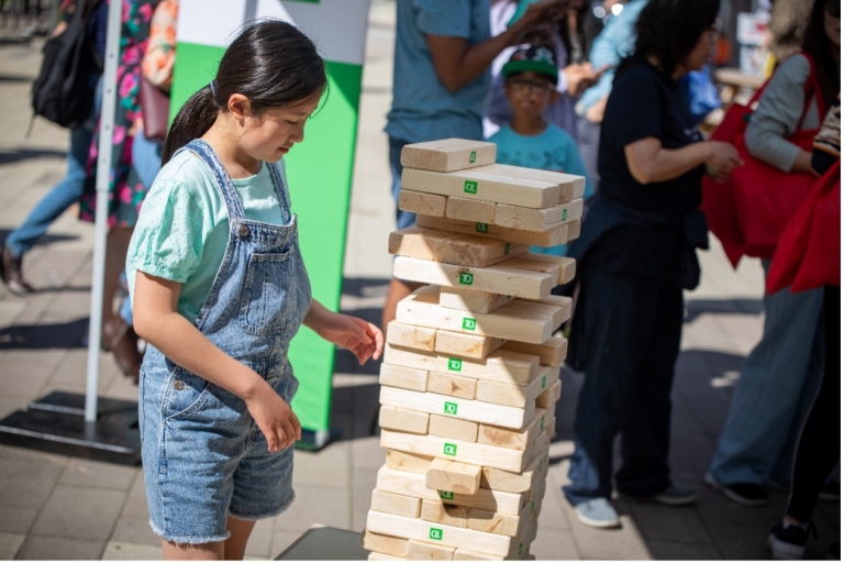 Girl plays jenga with a stack of blocks nearly as tall as she is. 